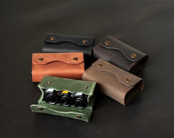 May include: A collection of five rectangular leather cases with snap closures, including black, tan, dark brown, and green options. One green case is open, revealing three cylindrical film canisters stored inside the main compartment.