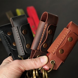 May include: A hand holding several leather keychains in various shades of brown and black. Each keychain has a loop for keys, secured with brass-colored rivets and stitching. The background is blurred, with hints of other colors.
