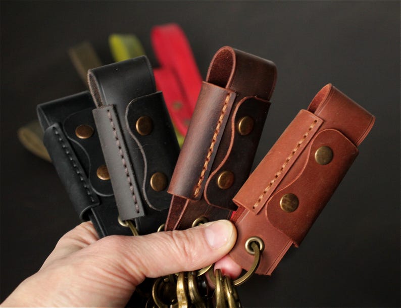 May include: A hand holding several leather keychains in various shades of brown and black. Each keychain has a loop for keys, secured with brass-colored rivets and stitching. The background is blurred, with hints of other colors.