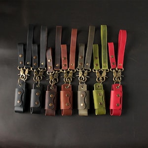 May include: A collection of leather keychains in various colors, including black, brown, green, and red. Each keychain features a leather holder with a snap closure and a brass-colored clasp and strap. The keychains are arranged against a dark background.