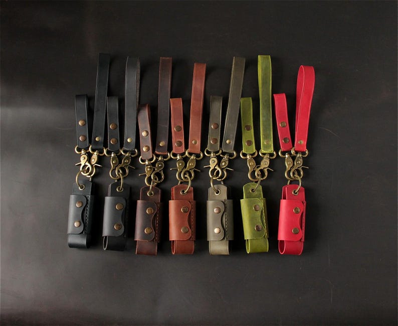 May include: A collection of leather keychains in various colors, including black, brown, green, and red. Each keychain features a leather holder with a snap closure and a brass-colored clasp and strap. The keychains are arranged against a dark background.