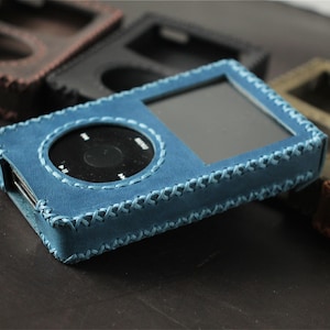 May include: A blue leather protective case for a digital music player, featuring a rectangular screen cutout and a circular click wheel opening, secured with visible cross-stitching along the edges, surrounded by other cases in various colors.