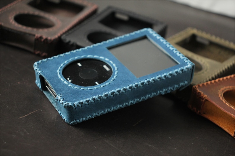 May include: A blue leather protective case for a digital music player, featuring a rectangular screen cutout and a circular click wheel opening, secured with visible cross-stitching along the edges, surrounded by other cases in various colors.
