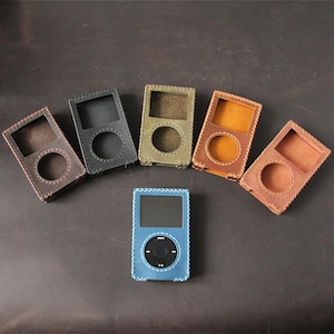 May include: Five leather cases for portable media players arranged in a semi-circle, featuring colors of dark brown, black, olive green, mustard yellow, and tan. A blue leather case containing a portable media player sits in the center.