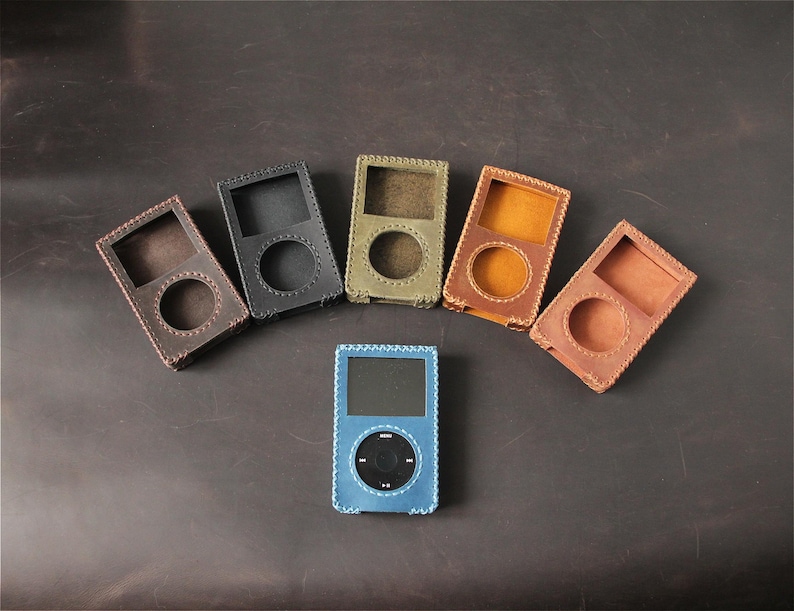 May include: Five leather cases for portable media players arranged in a semi-circle, featuring colors of dark brown, black, olive green, mustard yellow, and tan. A blue leather case containing a portable media player sits in the center.
