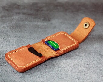 May include: A tan leather card holder with visible light-colored stitching and a brass snap button closure. The open case features two interior slots, one containing a blue and green card and the other holding a black card.