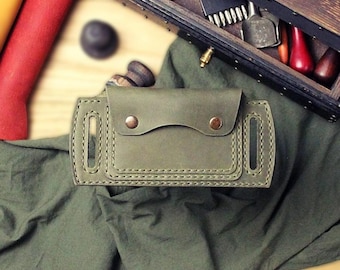 May include: An olive green leather belt sheath featuring a rectangular flap secured by two copper-toned metal snaps. The sheath has vertical slots on each side for belt attachment and is constructed with visible heavy-duty stitching along the edges.