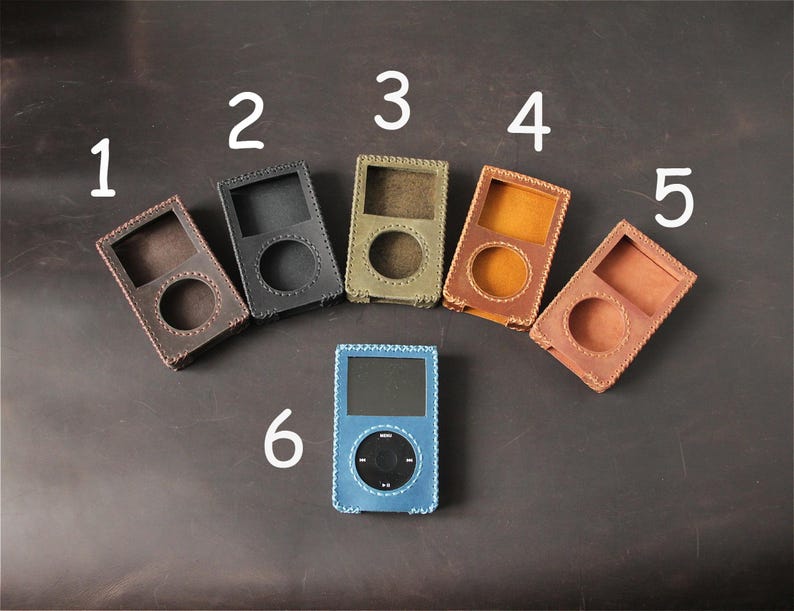 May include: Six leather cases for portable media players arranged in an arc. Cases 1 through 5 are empty, featuring stitched edges and cutouts for screens and click wheels. Case 6 is blue, contains a black media player, and is labeled with a 6.
