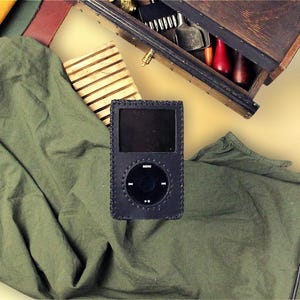 May include: A black leather iPod case with a stitched border, featuring a circular control pad. The case rests on a green fabric, near a wooden box filled with tools and leather scraps. The overall aesthetic suggests a handcrafted item.