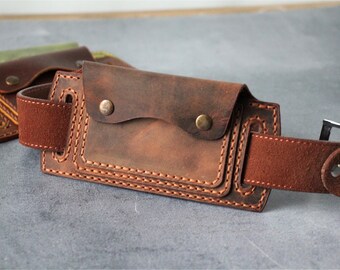 May include: Close-up of a brown leather belt pouch with a flap closure secured by two brass-colored rivets. The pouch is stitched with a contrasting thread and has a belt loop. Another similar pouch is visible in the background.