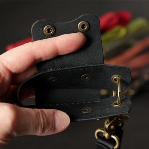 May include: A black leather phone case with brass-colored hardware. The case is held open, revealing the interior. The background is blurred, with hints of red, yellow, and black.
