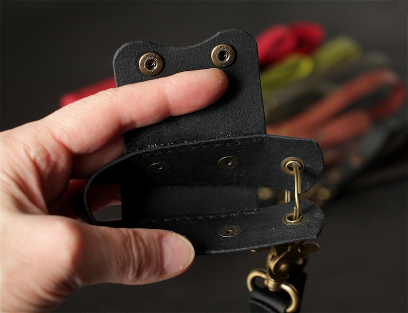 May include: A black leather phone case with brass-colored hardware. The case is held open, revealing the interior. The background is blurred, with hints of red, yellow, and black.