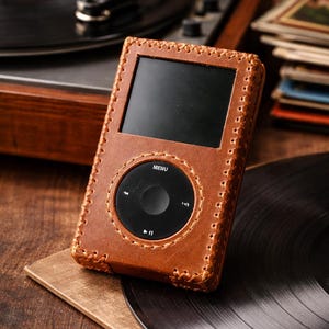May include: A brown leather case protects a black media player with a screen and control buttons. The case is stitched around the edges. A vintage record player and vinyl records are in the background, suggesting a retro theme.