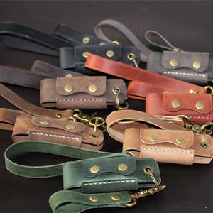 May include: A collection of handcrafted leather keychains in various colors, including brown, green, and black. Each keychain features a small pouch with a snap closure and a brass-colored clasp for attaching keys. The keychains have a loop for easy carrying.