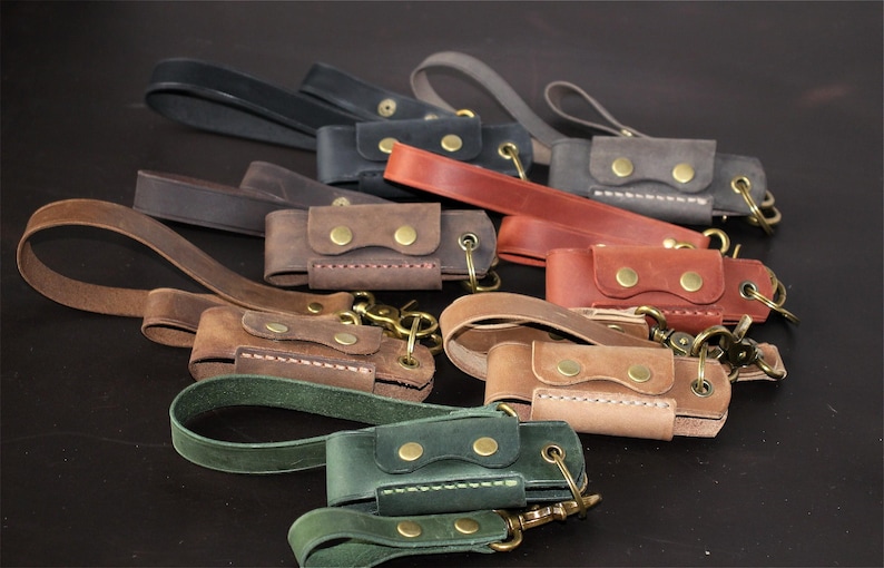 May include: A collection of handcrafted leather keychains in various colors, including brown, green, and black. Each keychain features a small pouch with a snap closure and a brass-colored clasp for attaching keys. The keychains have a loop for easy carrying.
