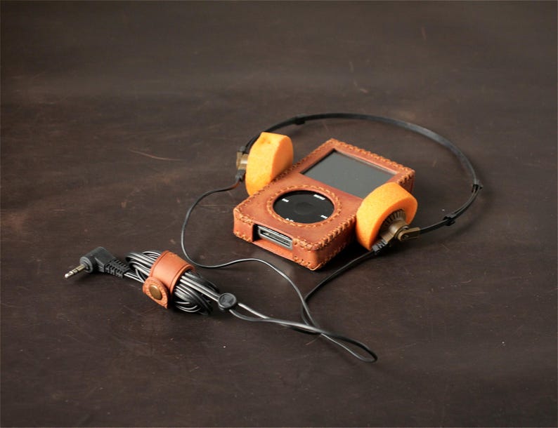 May include: A digital music player encased in a brown leather cover with visible stitching, connected to black wired headphones featuring bright orange foam ear pads. A small brown leather cable organizer with a metal snap button secures the black cord.