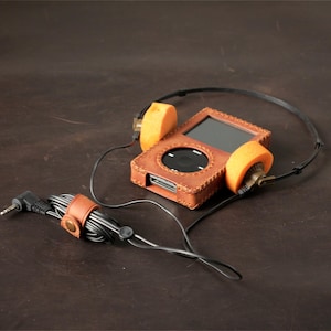 May include: A digital music player encased in a tan leather cover with visible stitching, connected to black wired headphones featuring bright orange foam ear pads. A small tan leather cable organizer with a metal snap button secures the black cord.