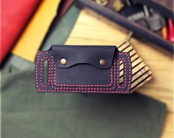 May include: A dark navy blue leather belt sheath featuring a flap closure secured with two brass-colored metal snaps. The rectangular leather piece has vertical slots on each side and is stitched with thick, contrasting red thread.