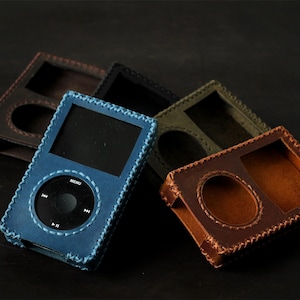 May include: A collection of rectangular leather cases for portable media players, featuring hand-stitched edges. The cases are available in blue, dark brown, black, and olive green, each with a cutout for the screen and circular click wheel.