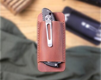 May include: A brown leather knife sheath with a stainless steel pocket clip, holding a black and silver folding knife. The sheath is stitched and designed to hold the knife securely. The background includes a wooden surface and a dark object.