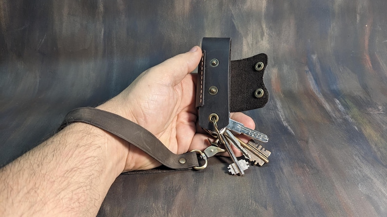 May include: A black leather key holder with a wrist strap, holding several keys. The key holder has a riveted design and a metal clasp. The wrist strap is brown.