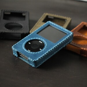 May include: A blue leather protective case for a digital music player, featuring visible stitched edges and a cutout for the circular control wheel and screen. Several other cases in black, brown, and olive green are arranged in the background.