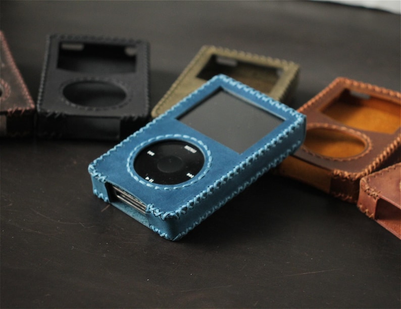 May include: A blue leather protective case for a digital music player, featuring visible stitched edges and a cutout for the circular control wheel and screen. Several other cases in black, brown, and olive green are arranged in the background.