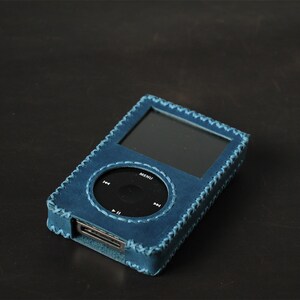 May include: A blue leather protective case for a digital music player. The case features a rectangular cutout for the screen and a circular cutout for the click wheel, with visible cross-stitching along the edges and an opening at the bottom.