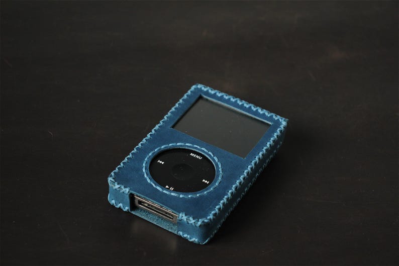 May include: A blue leather protective case for a digital music player. The case features a rectangular cutout for the screen and a circular cutout for the click wheel, with visible cross-stitching along the edges and an opening at the bottom.