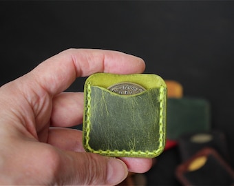 May include: A square olive green leather coin pouch featuring a curved top opening and bright yellow contrast stitching along the edges. A metallic coin with intricate embossed detailing is partially visible inside the small pocket.