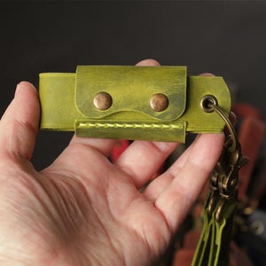 May include: A lime green leather key holder featuring a rectangular flap secured by two antique brass-colored metal snaps. The accessory includes a matching lime green stitched edge and a brass-toned metal eyelet with a swivel lobster clasp attached.