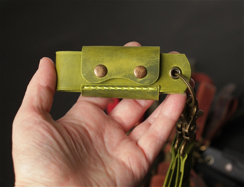May include: A lime green leather key holder featuring a rectangular flap secured by two antique brass-colored metal snaps. The accessory includes a matching lime green stitched edge and a brass-toned metal eyelet with a swivel lobster clasp attached.