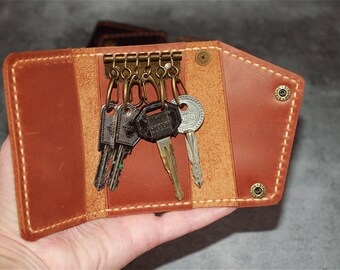 May include: A brown leather key holder with a brass key ring and several keys attached. The key holder is open, revealing the keys. The leather has a stitched border and brass rivets.
