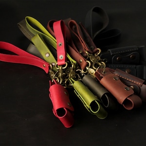May include: A collection of leather keychains in various colors, including red, green, brown, and black. Each keychain features a wrist strap, a metal clasp, and a small leather pouch. The keychains are arranged on a dark surface.
