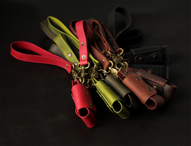 May include: A collection of leather keychains in various colors, including red, green, brown, and black. Each keychain features a wrist strap, a metal clasp, and a small leather pouch. The keychains are arranged on a dark surface.