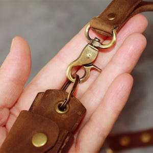 May include: Close-up of a brown leather key fob with a brass-colored clasp and hardware. The key fob is attached to a matching leather strap. The leather appears to be distressed, with visible grain and texture.