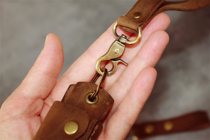 May include: Close-up of a brown leather key fob with a brass-colored clasp and hardware. The key fob is attached to a matching leather strap. The leather appears to be distressed, with visible grain and texture.