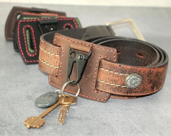 May include: A brown leather belt with a metal buckle and a decorative silver button. A black metal carabiner clip holds a set of keys. Several other leather belt accessories are visible in the background.