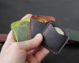 May include: A collection of small, square leather pouches featuring visible hand-stitched edges. The pouches are available in various colors including lime green, dark brown, and black, each designed to hold a single round coin or token.