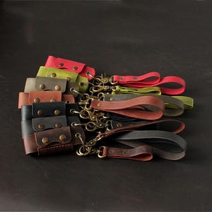 May include: A collection of leather wrist straps in various colors, including red, green, brown, and black. Each strap features a brass-colored clasp and a snap closure. The straps are arranged on a dark surface.