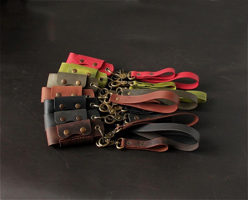 May include: A collection of leather wrist straps in various colors, including red, green, brown, and black. Each strap features a brass-colored clasp and a snap closure. The straps are arranged on a dark surface.