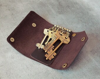 May include: A dark brown leather key holder featuring a metal key ring mechanism with multiple attached keys. The holder is secured with brass-colored snap buttons and displays the word CLASS embossed on one of the metal keys.