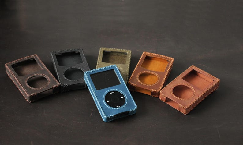 May include: A collection of six rectangular leather cases for portable media players, each featuring a circular cutout for a click wheel and a square cutout for a screen. The cases are available in blue, black, olive green, and various shades of brown.