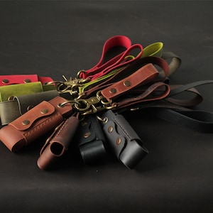 May include: A collection of leather keychains in various colors, including red, green, brown, and black. Each keychain features a leather case with a snap closure and a metal clasp. The keychains are arranged on a dark surface.