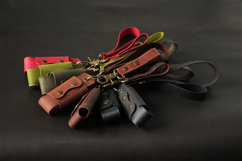 May include: A collection of leather keychains in various colors, including red, green, brown, and black. Each keychain features a leather case with a snap closure and a metal clasp. The keychains are arranged on a dark surface.