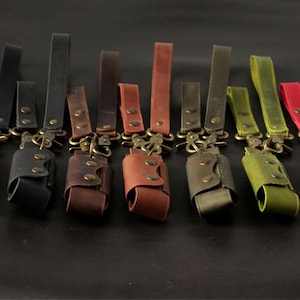 May include: A collection of leather keychains in various colors, including black, brown, and red. Each keychain features a leather pouch with a snap closure and a brass-colored clasp and loop. The keychains are arranged on a dark surface.