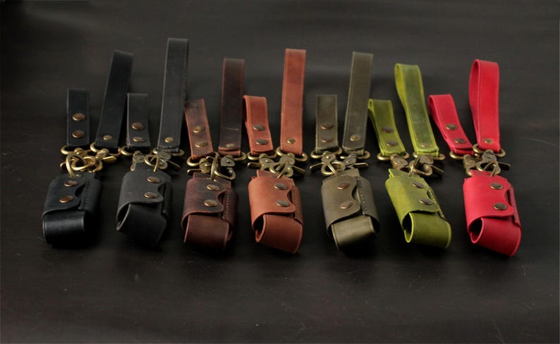 May include: A collection of leather keychains in various colors, including black, brown, and red. Each keychain features a leather pouch with a snap closure and a brass-colored clasp and loop. The keychains are arranged on a dark surface.