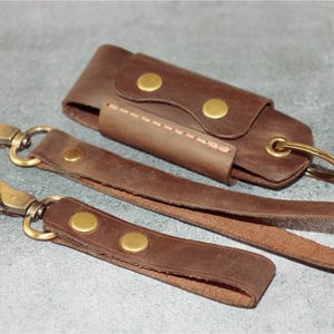 May include: A set of brown leather keychains and a key holder. The keychains have brass-colored clasps and rivets. The key holder is a small, rectangular case with a snap closure. The items are arranged on a gray surface.