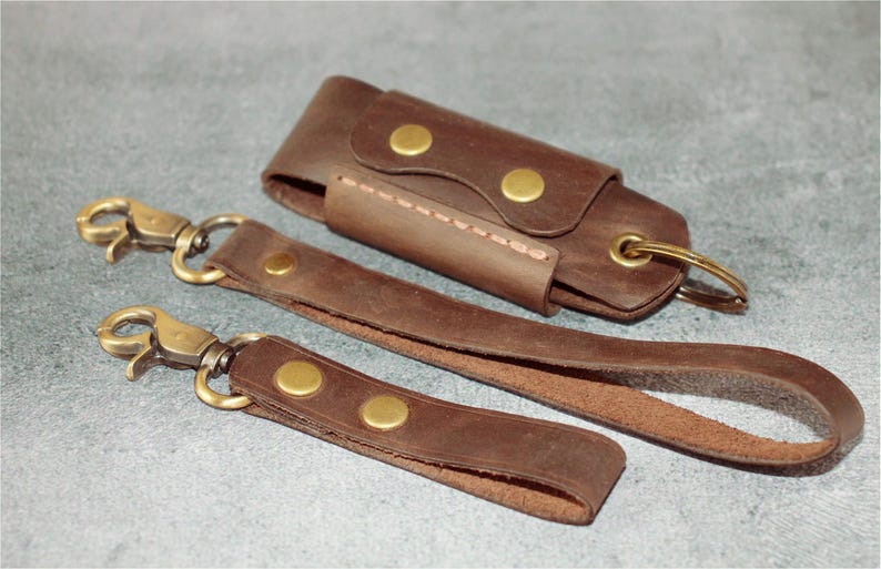 May include: A set of brown leather keychains and a key holder. The keychains have brass-colored clasps and rivets. The key holder is a small, rectangular case with a snap closure. The items are arranged on a gray surface.