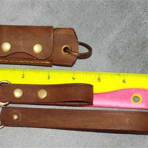 May include: Brown leather keychains and a key holder with brass hardware. The keychains have a swivel clasp and are secured with brass rivets. The key holder has a snap closure. A yellow and pink ruler is in the background.